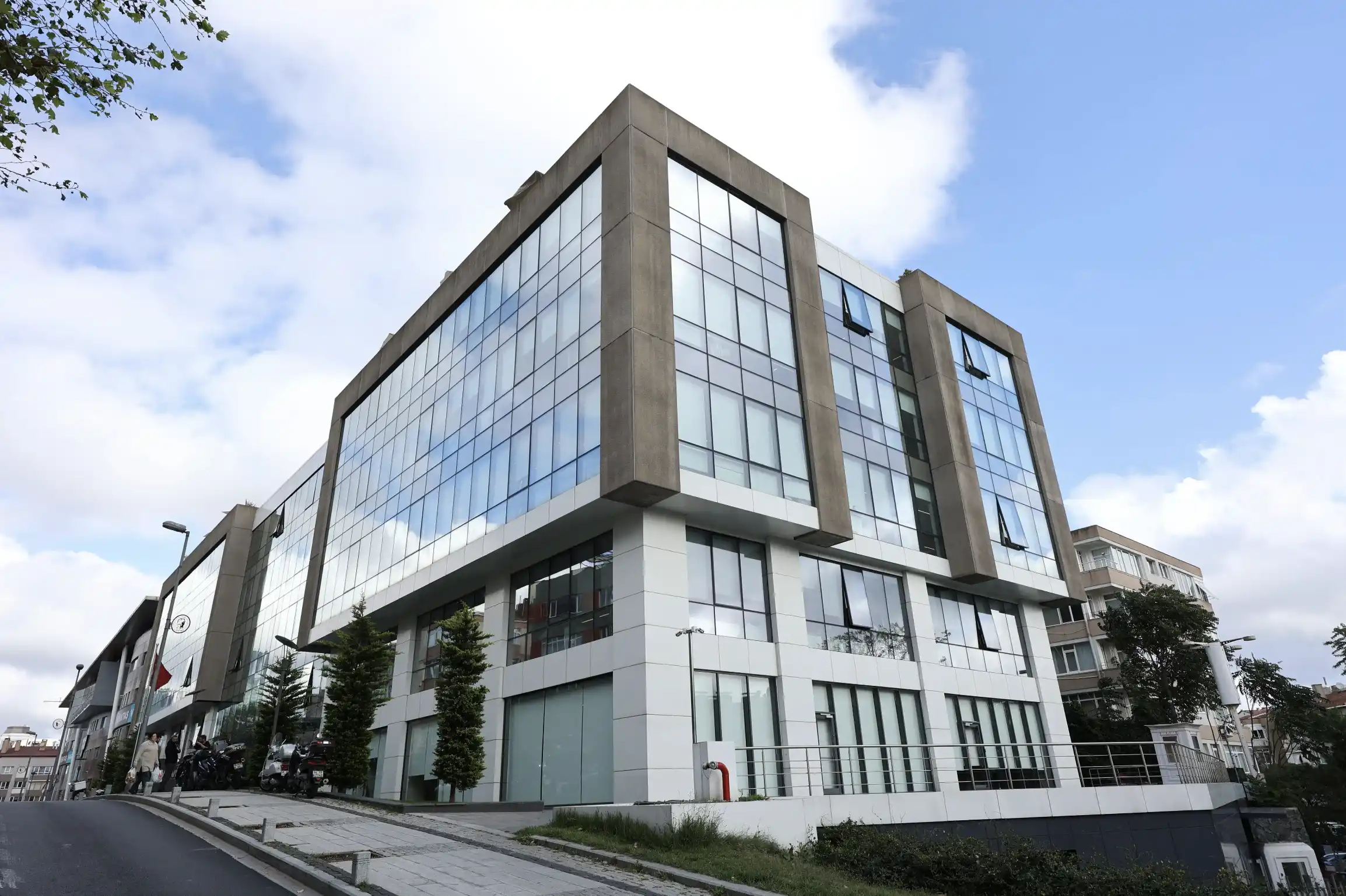 Modern MM Plaza office building where a chemical industry company is located, depicted under a bright sky with light cloud cover. The building features a contemporary design with a glass facade, vertical stone pillars, and a landscaped front area with greenery and trees. The image captures the professional and advanced setting of the company's headquarters, situated in an accessible urban area, emphasizing the industry presence and forward-thinking ethos of the company.